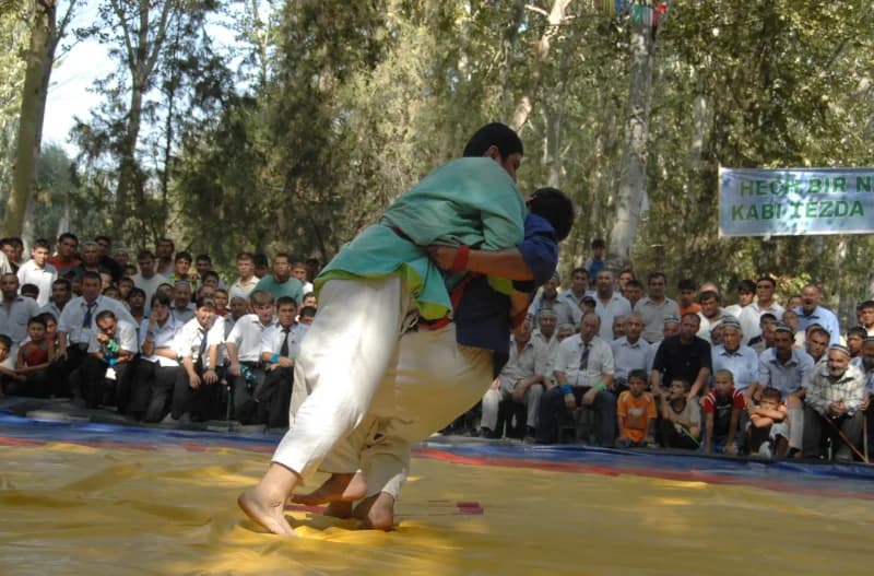 Competitors grapple during a traditional wrestling match on a yellow mat outdoors