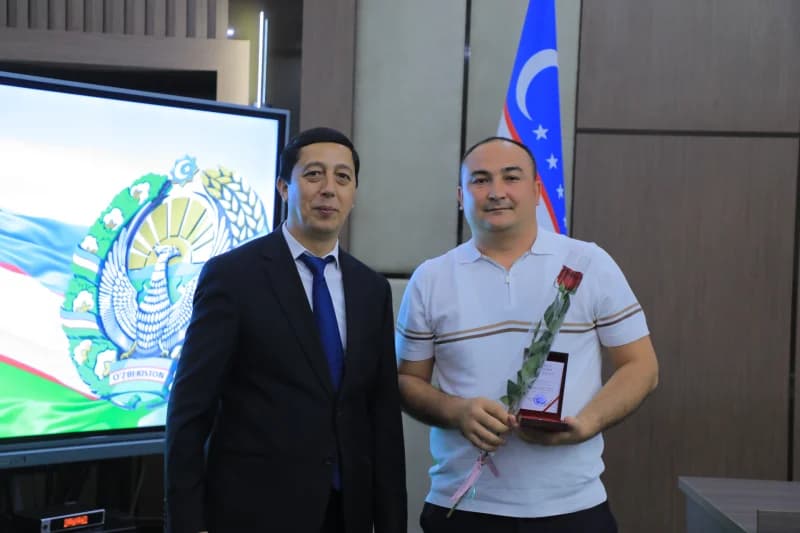 Two men pose indoors in front of a screen displaying a state emblem