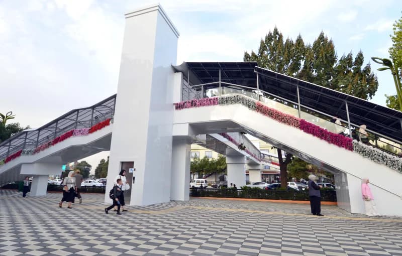 Pedestrians walk on a paved square near a modern overpass and elevator tower in an urban area