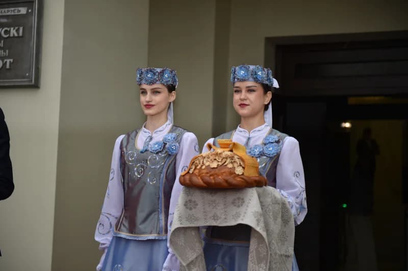 Anʼanaviy kiyimdagi ikki ayol bezatilgan nonni ushlab turibdi