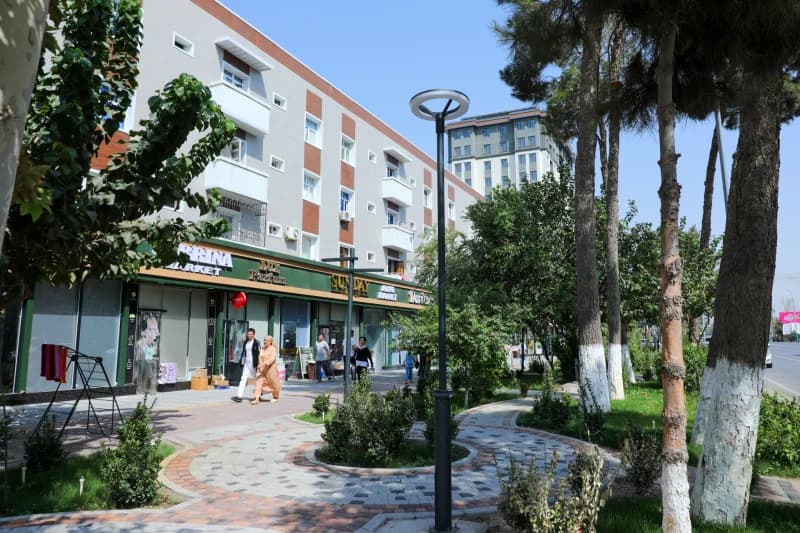People walking along a city sidewalk next to commercial storefronts and a paved park area
