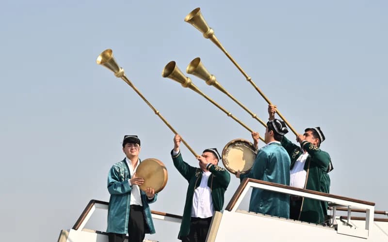 Performers in traditional clothing play long brass horns and frame drums