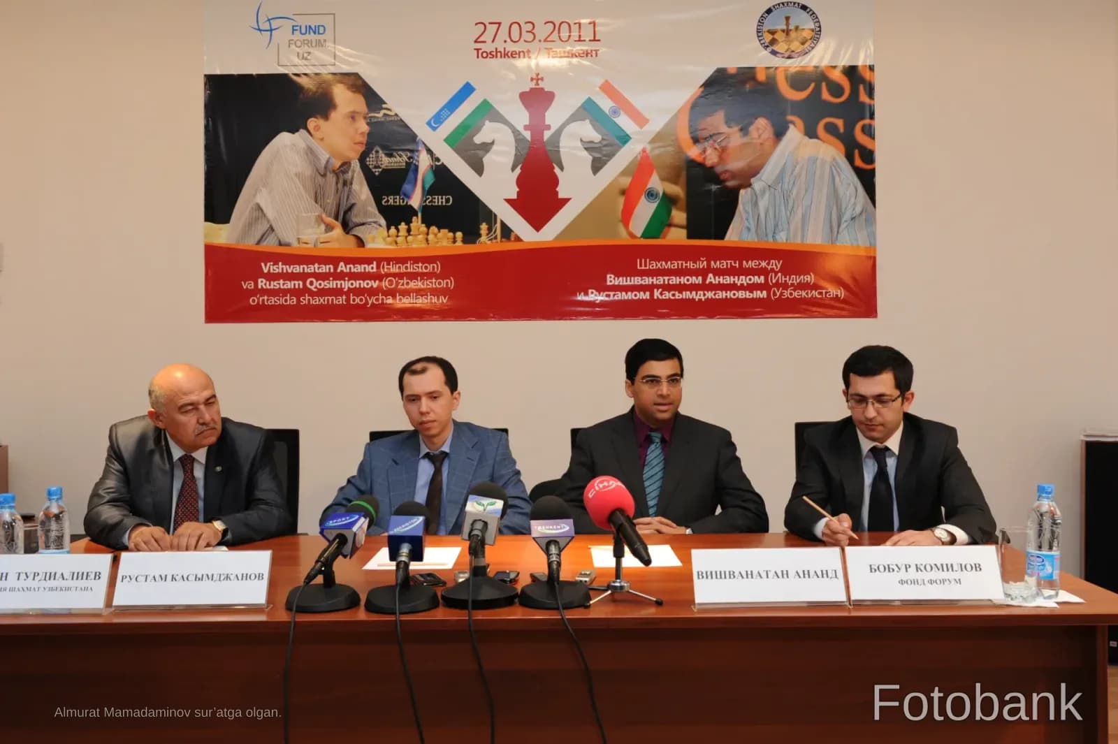 Four men seated at a press conference table with microphones and nameplates