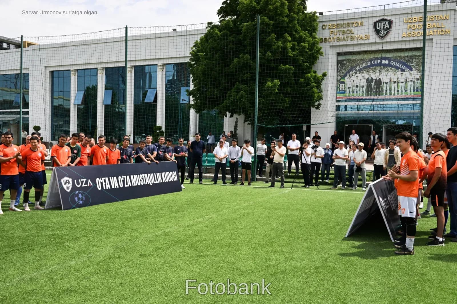Ochiq sunʼiy chimli futbol maydonida toʻplangan odamlar va oʻyinchilar guruhlari