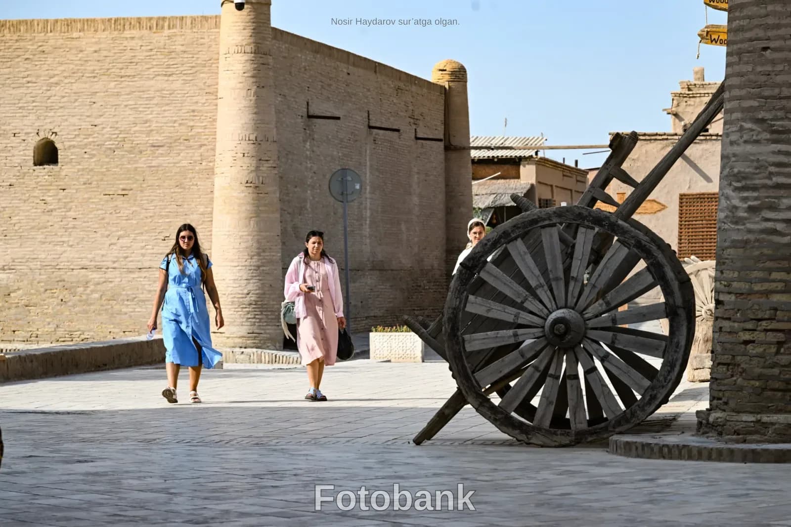 People walking past historic brick buildings and a large wooden cart wheel