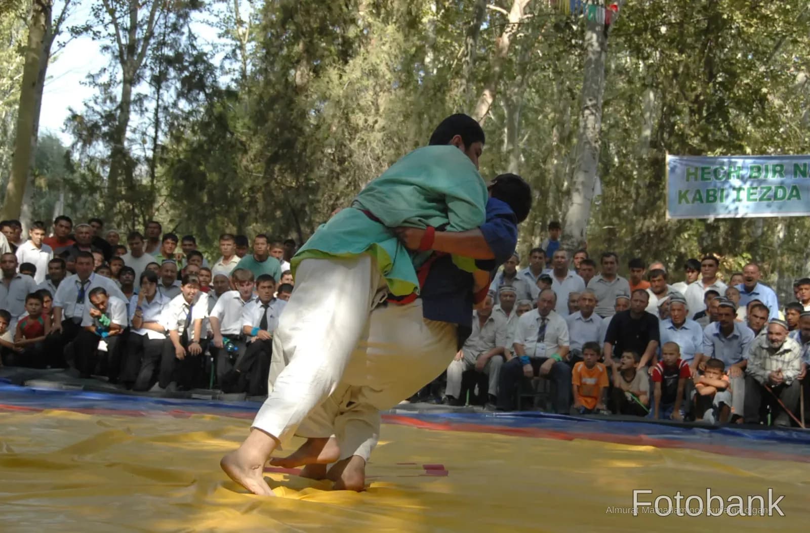 Competitors grapple during a traditional wrestling match on a yellow mat outdoors
