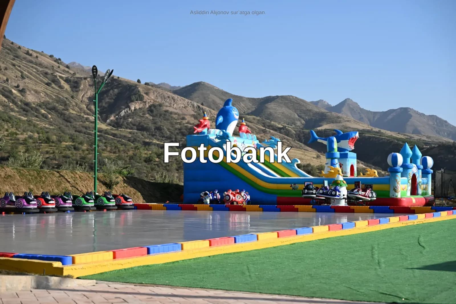Togʻ manzarasi fonida shishiriladigan sirpanchiq va bamperli avtomobillar bilan ochiq koʻngilochar park