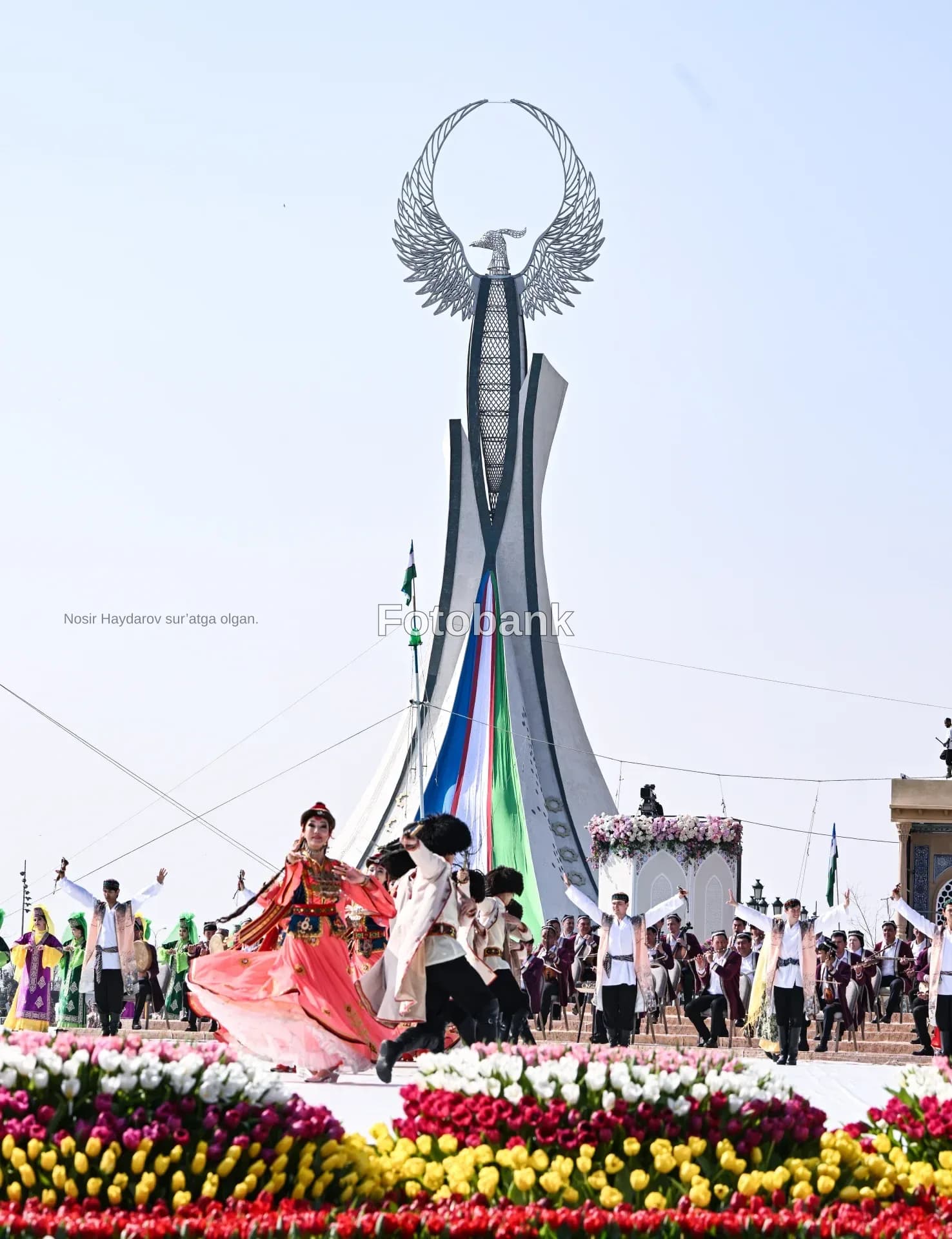 Performers in traditional clothing dancing outdoors near a large monument and flower displays