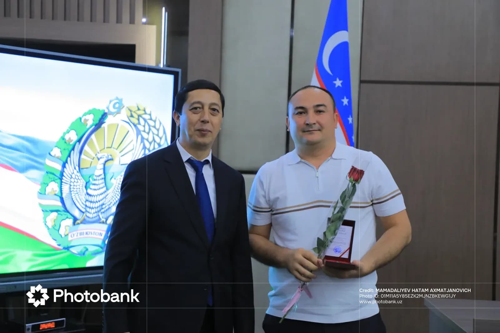 Ikki erkak bino ichida davlat gerbi aks ettirilgan ekran oldida suratga tushmoqda.