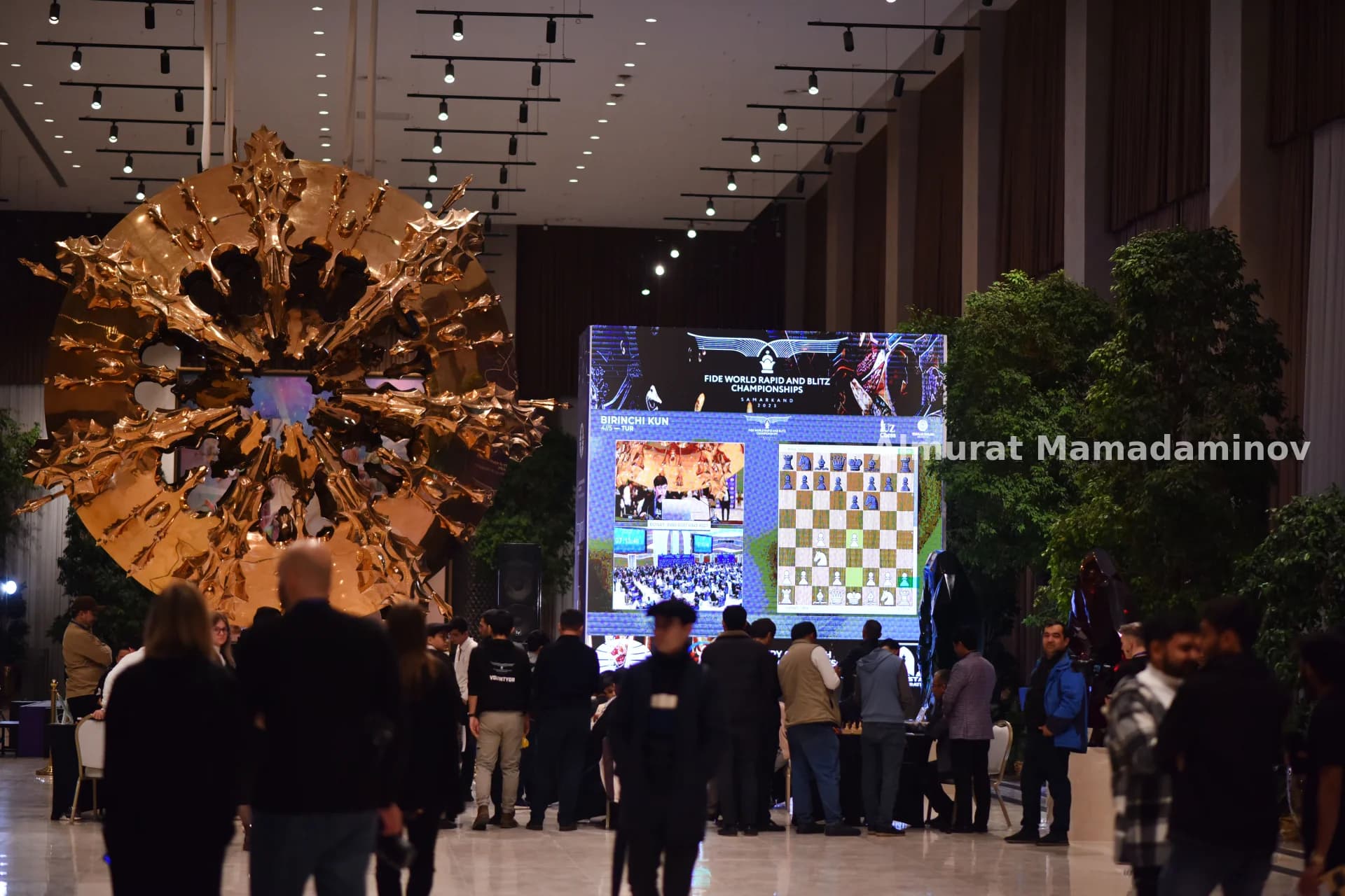 People gathered in a large hall with a screen displaying chess matches