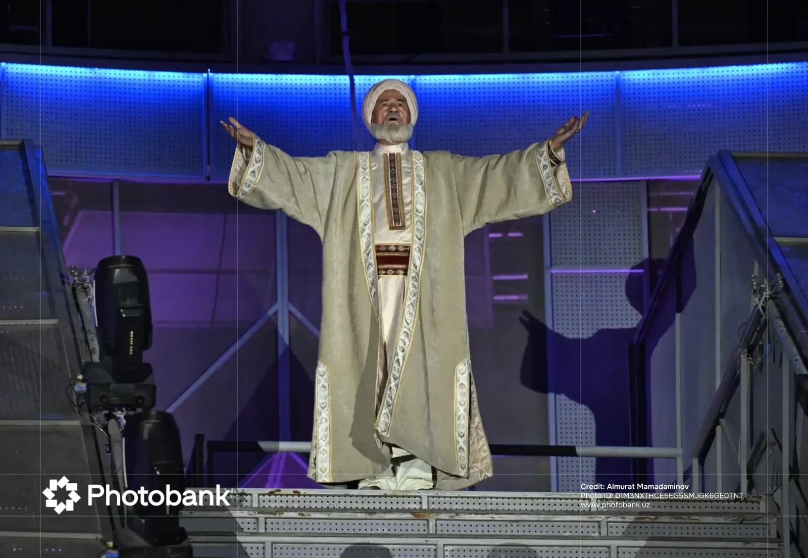 Elderly man in traditional attire standing with outstretched arms on stage