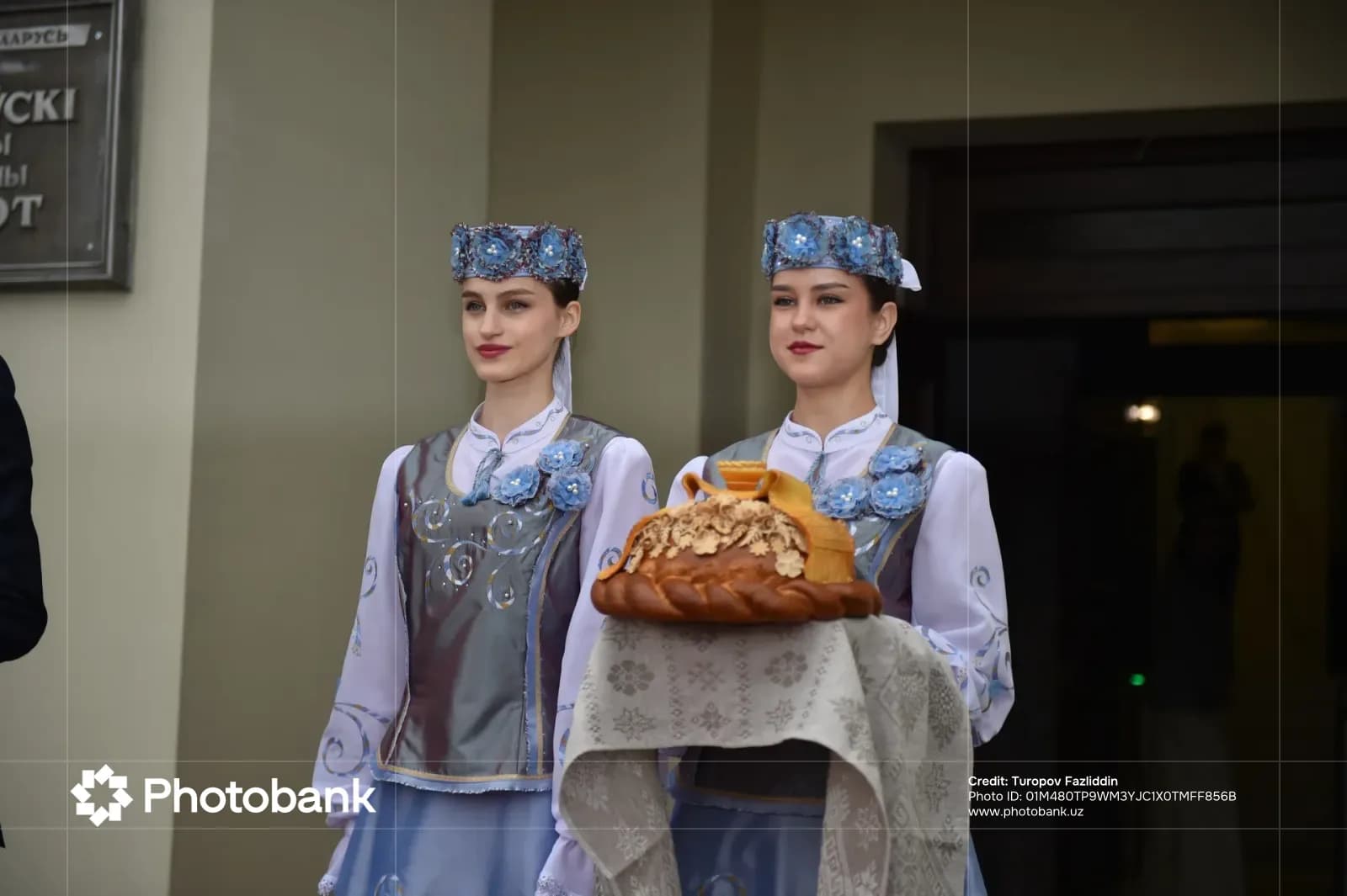 Anʼanaviy kiyimdagi ikki ayol bezatilgan nonni ushlab turibdi