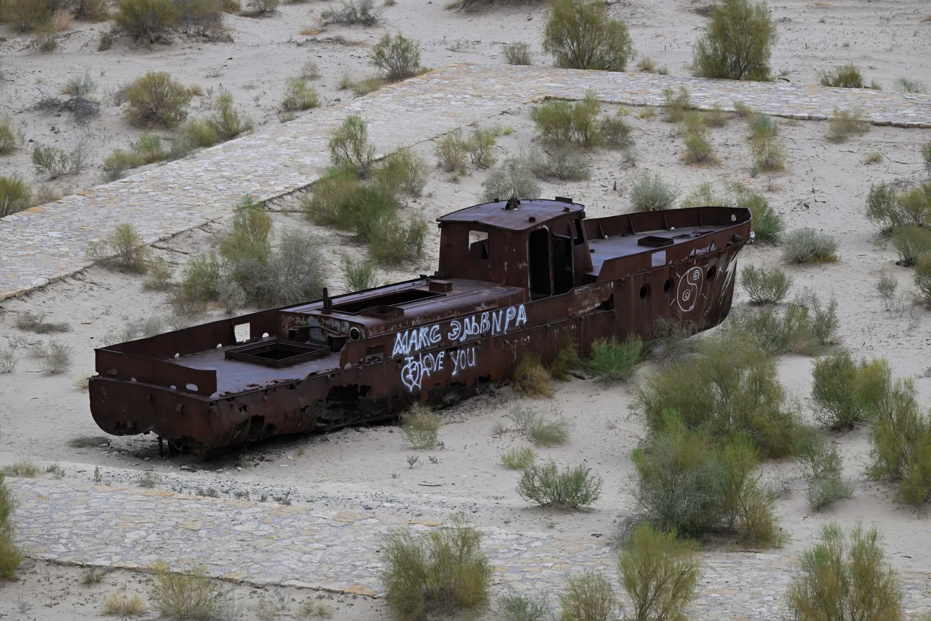 Rusted metal vessel resting on dry sand surrounded by sparse vegetation