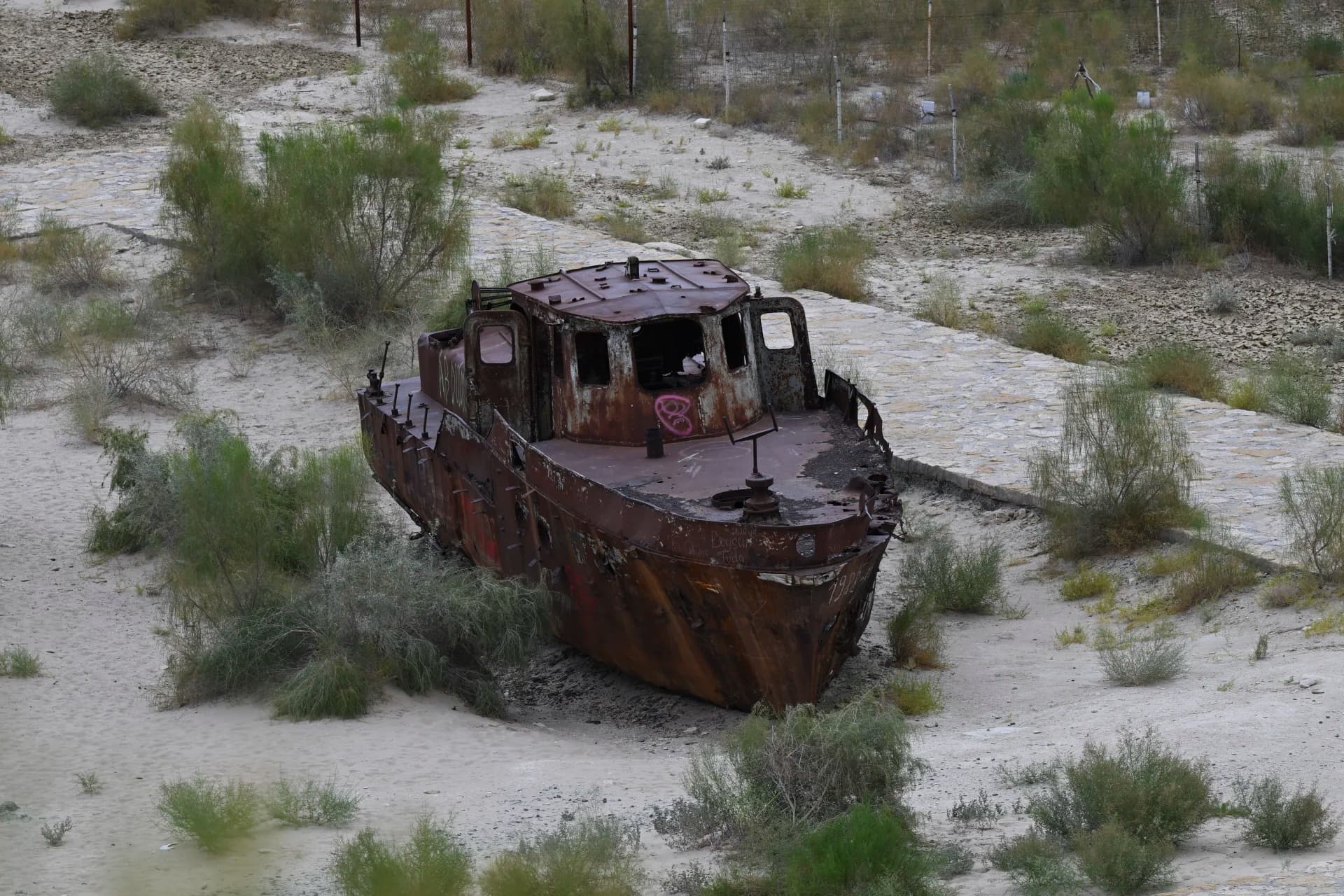 A rusted shipwreck stranded on dry, sandy terrain surrounded by sparse vegetation