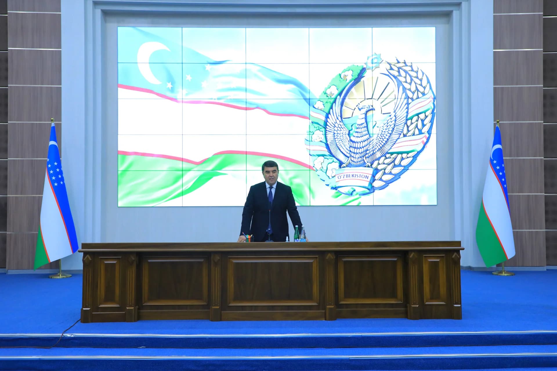 Человек, стоящий за большим деревянным столом перед дисплеем
