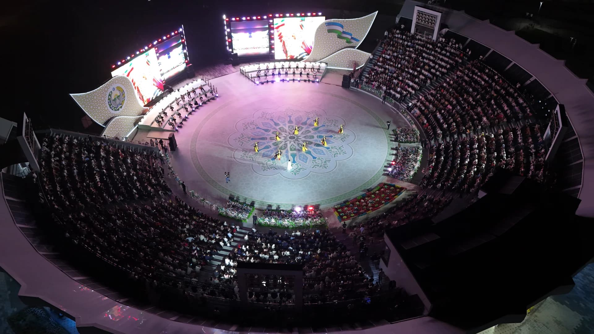 High-angle view of a nighttime arena event with performers on a central decorated stage surrounded by an audience