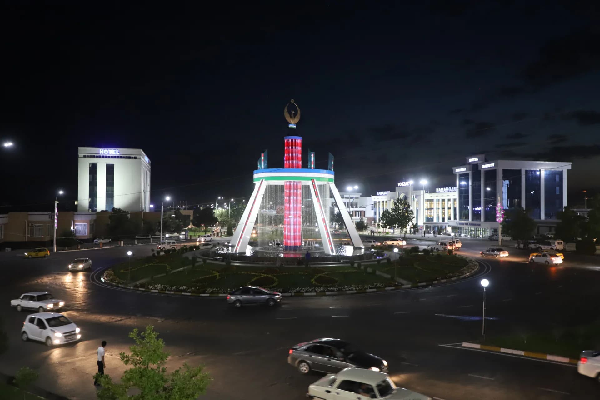Illuminated city square with central fountain and traffic at night