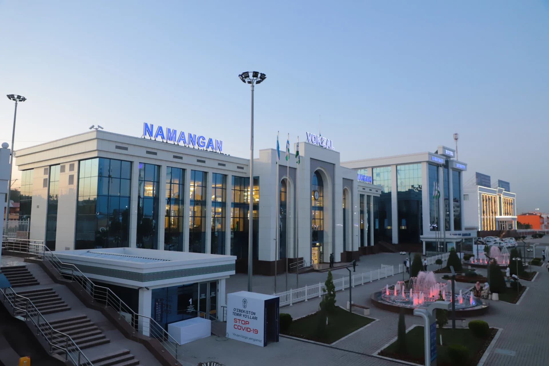 Exterior view of a modern railway station building with illuminated signs and a public square