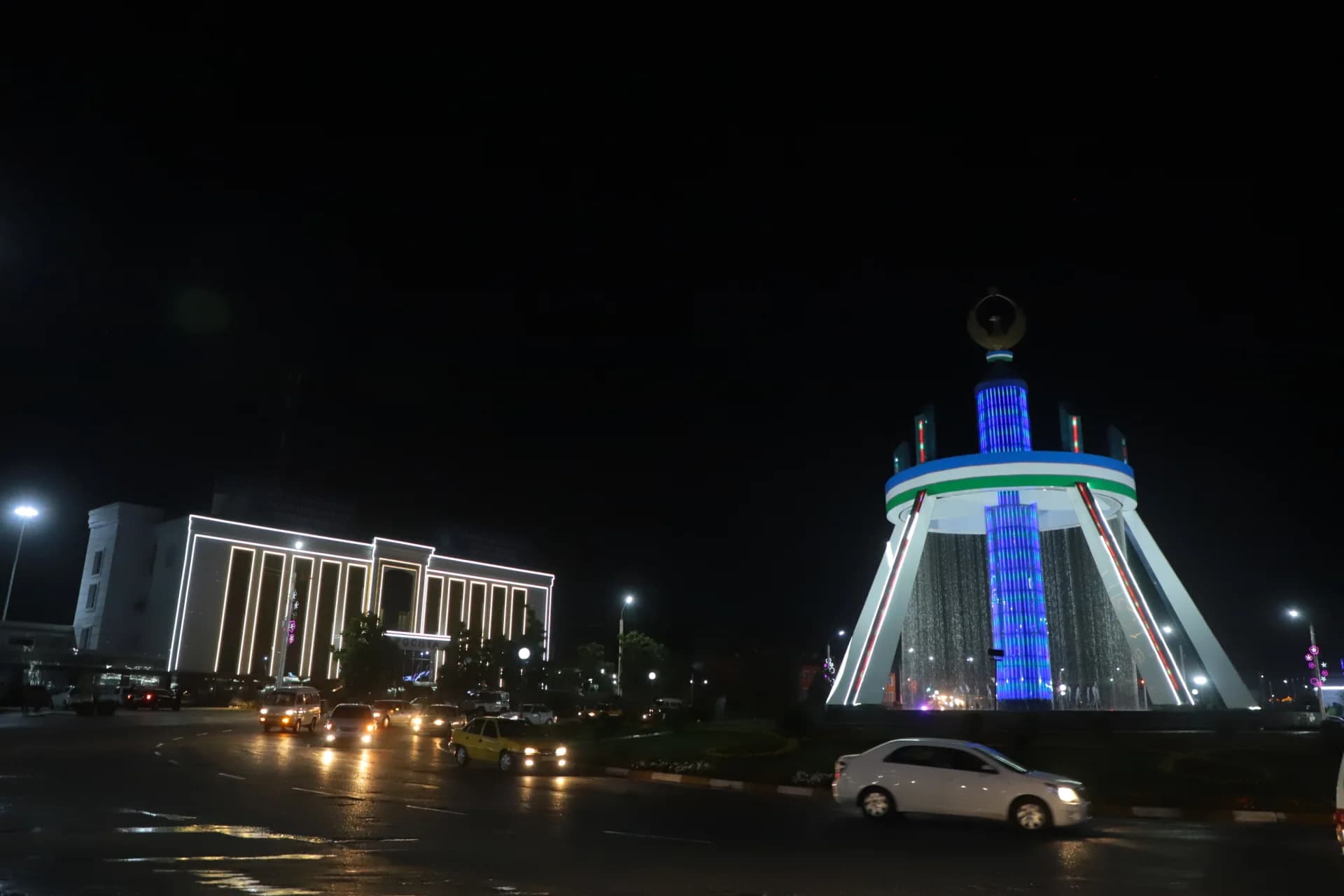 Night view of illuminated city square with fountain monument and vehicle traffic