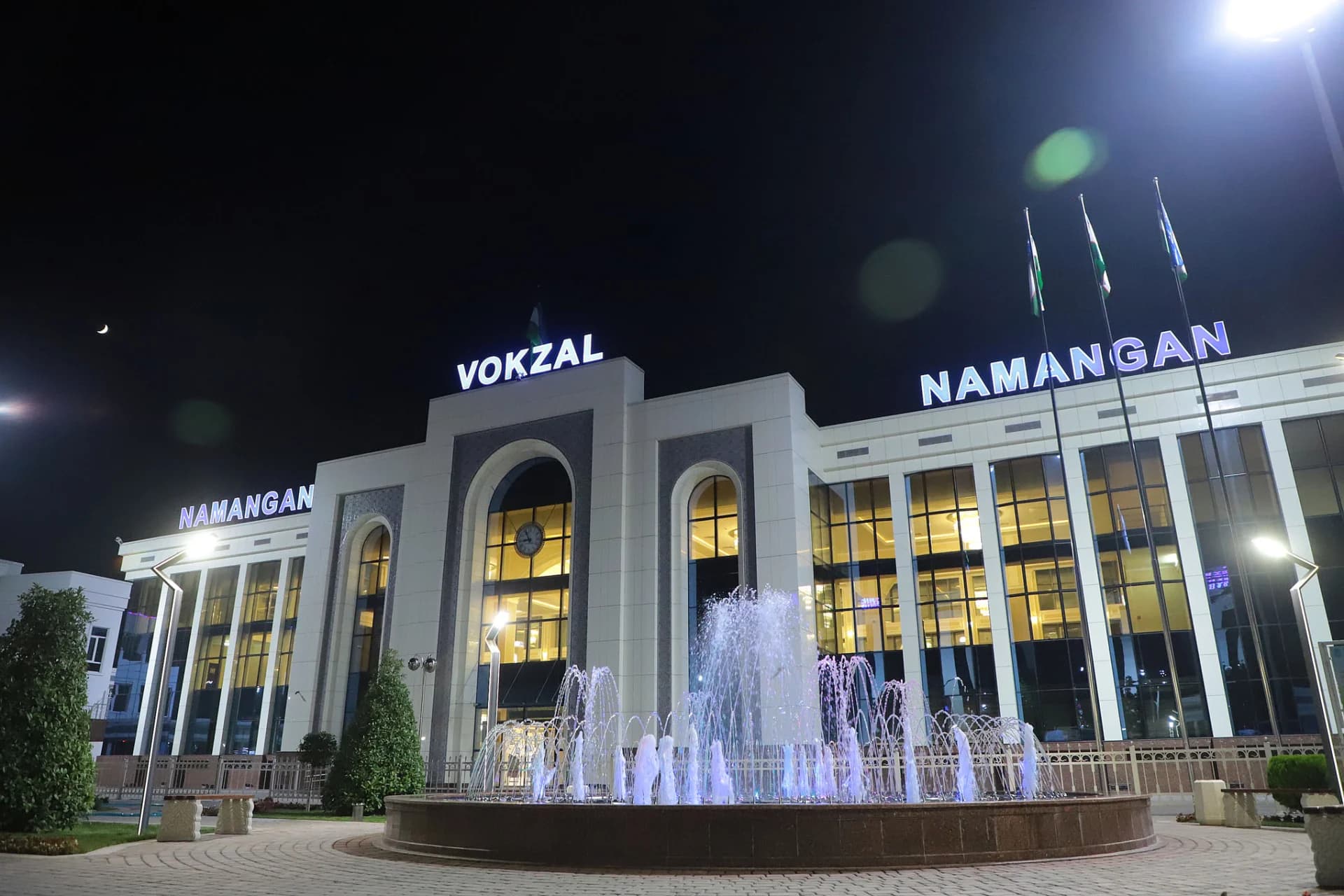 Illuminated railway station building at night with a fountain