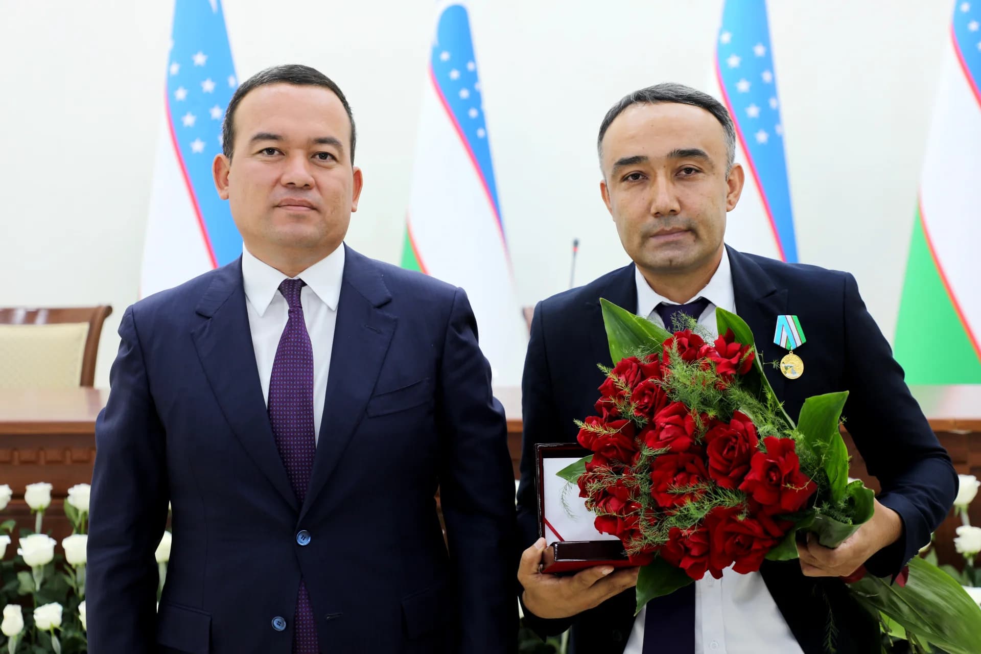 Two men pose indoors in front of flags