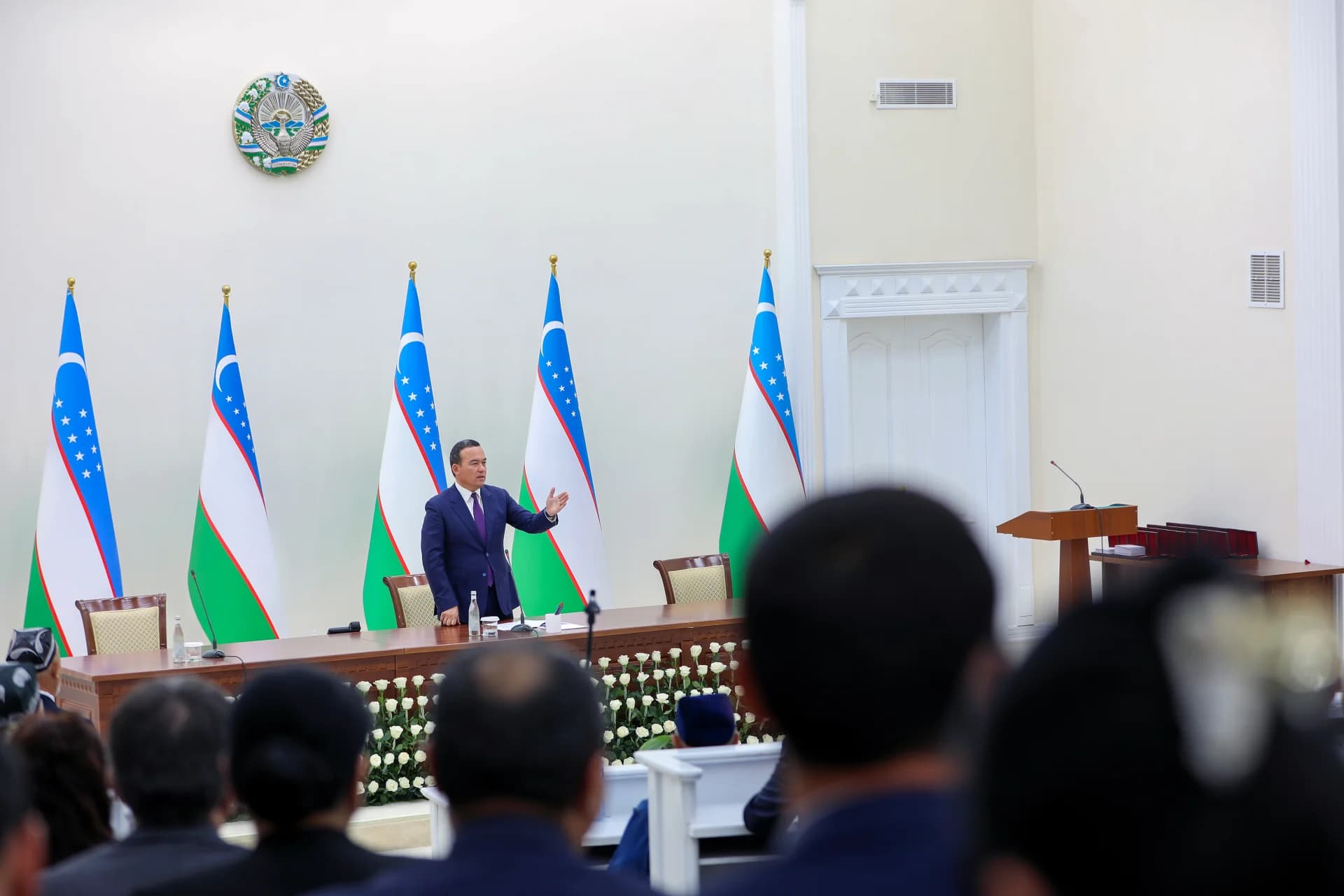 Person speaking before an audience in a hall with flags