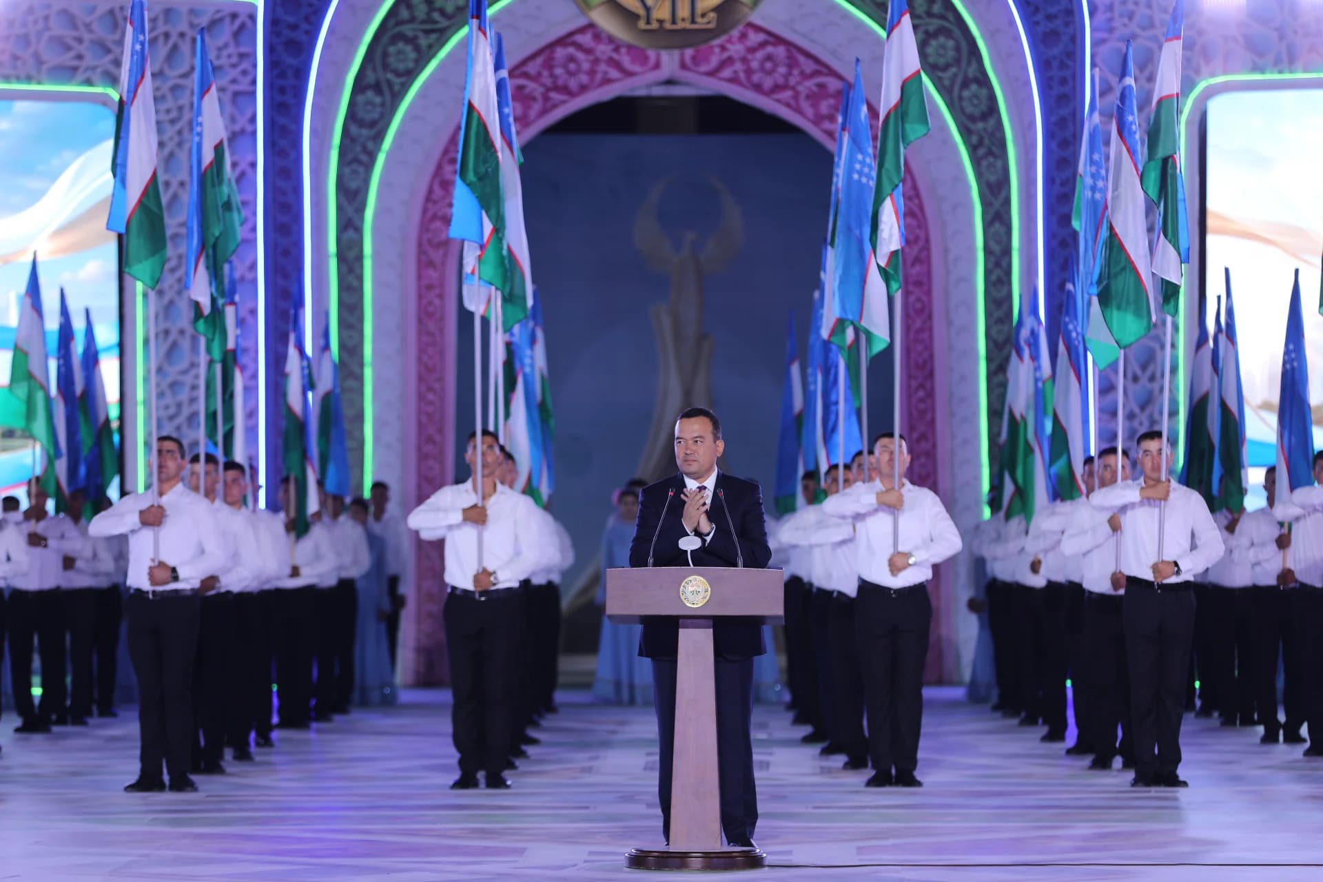 Man speaking at a podium before a gathering holding flags