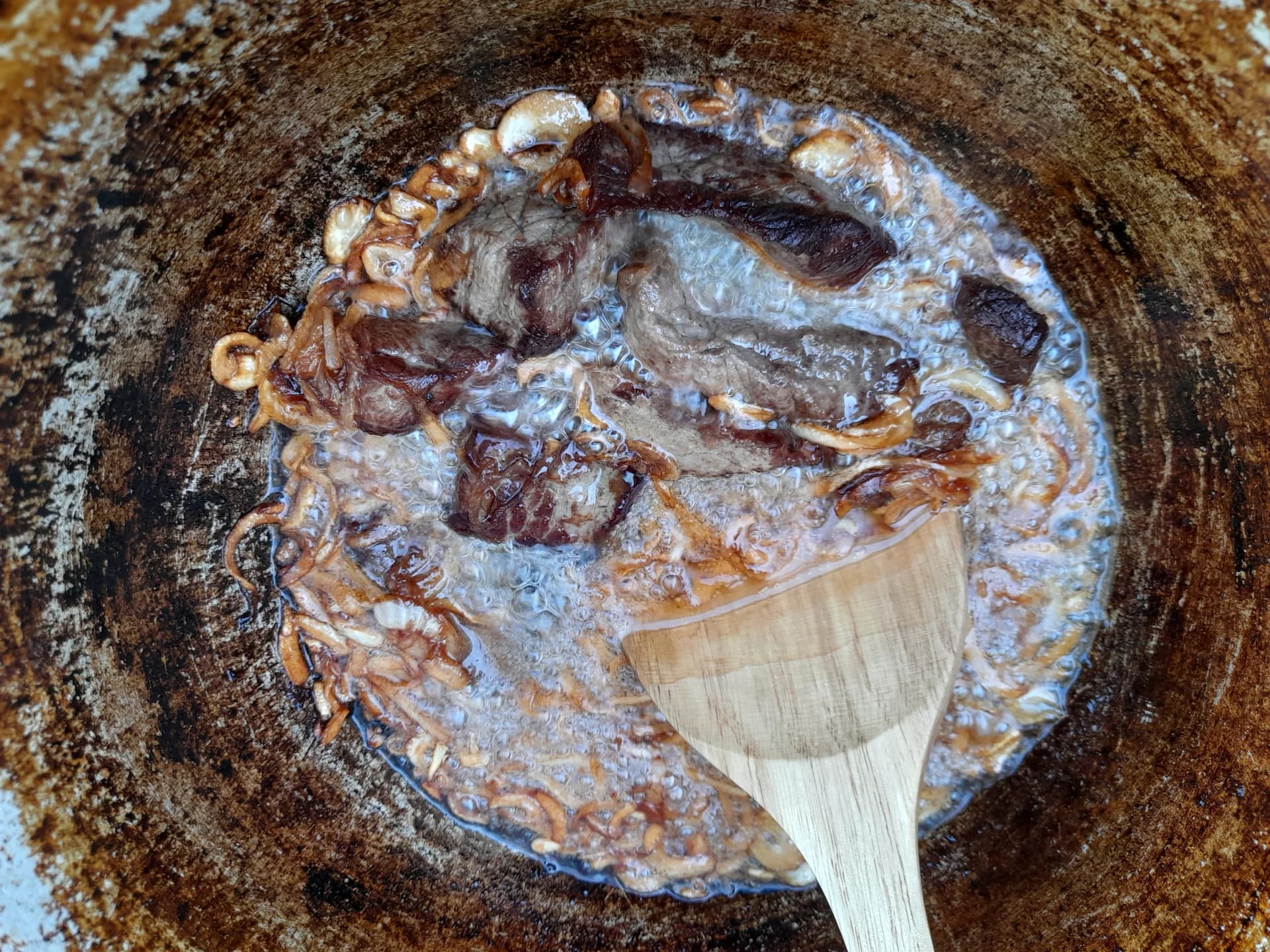 Жарка мяса и лука в глубокой металлической кастрюле с помощью деревянной ложки.