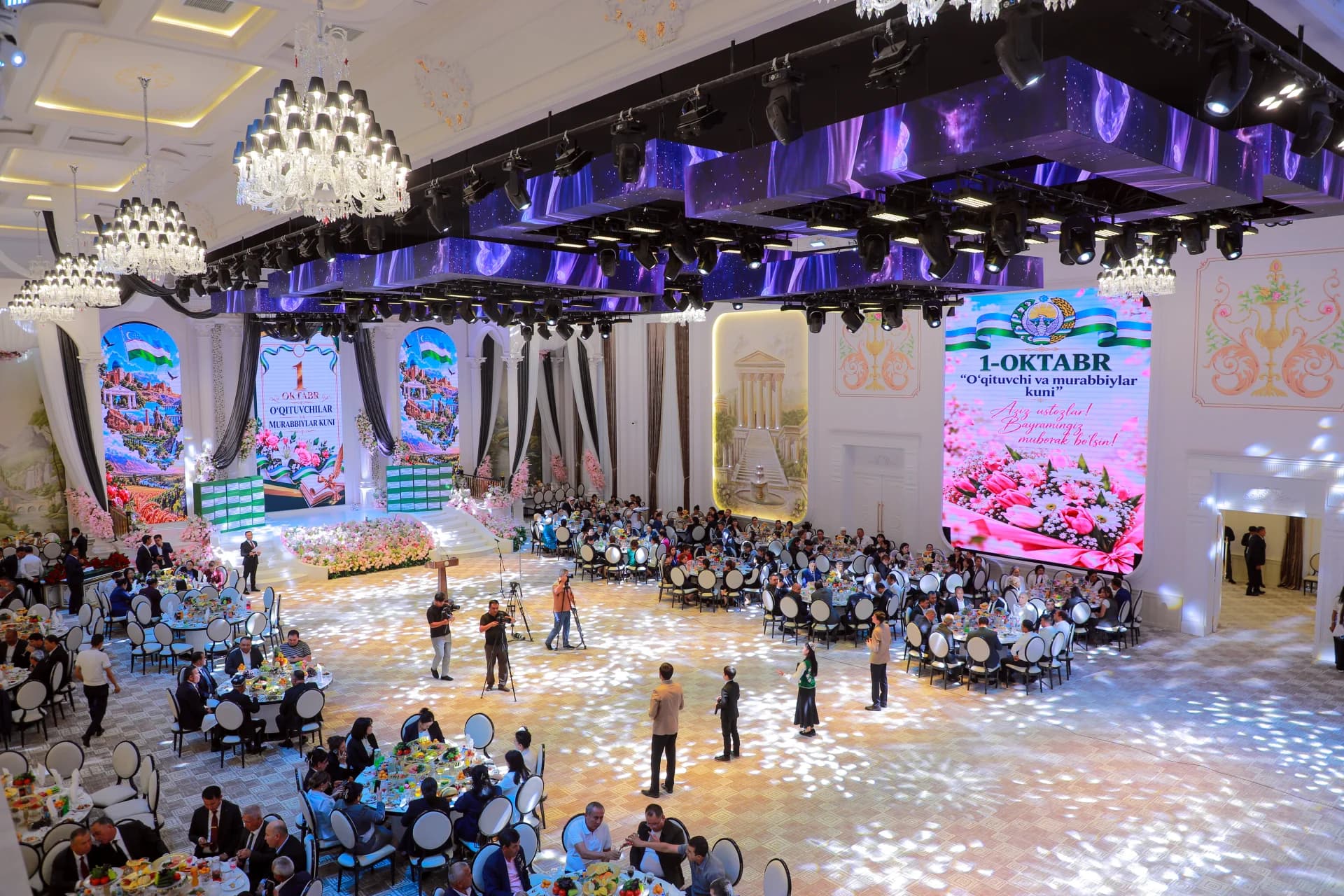 People gathered in a large decorated hall with large screens displaying text
