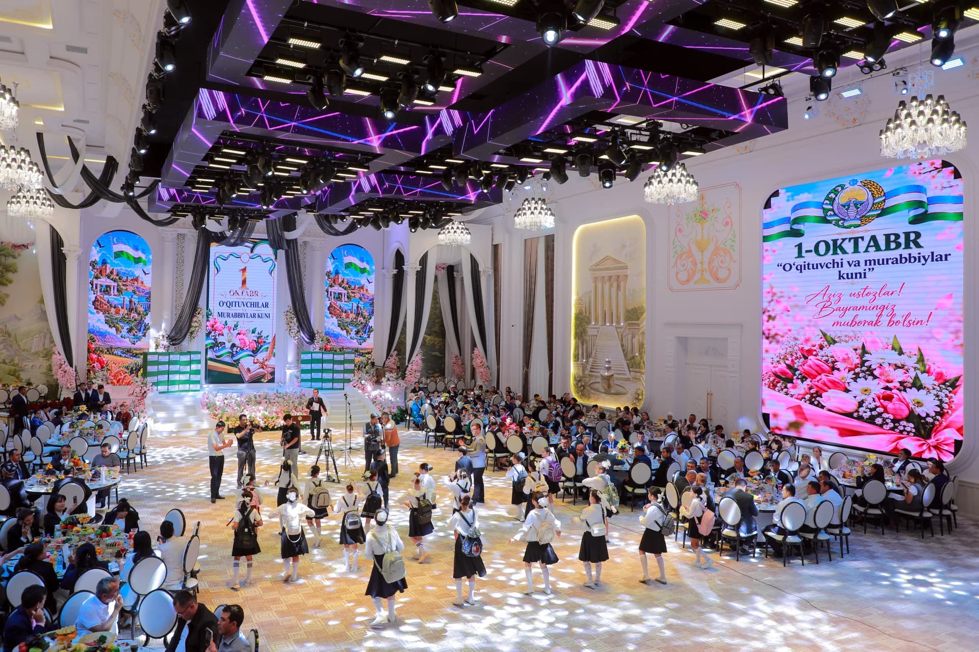 People gathered in a large decorated hall with large screens displaying text